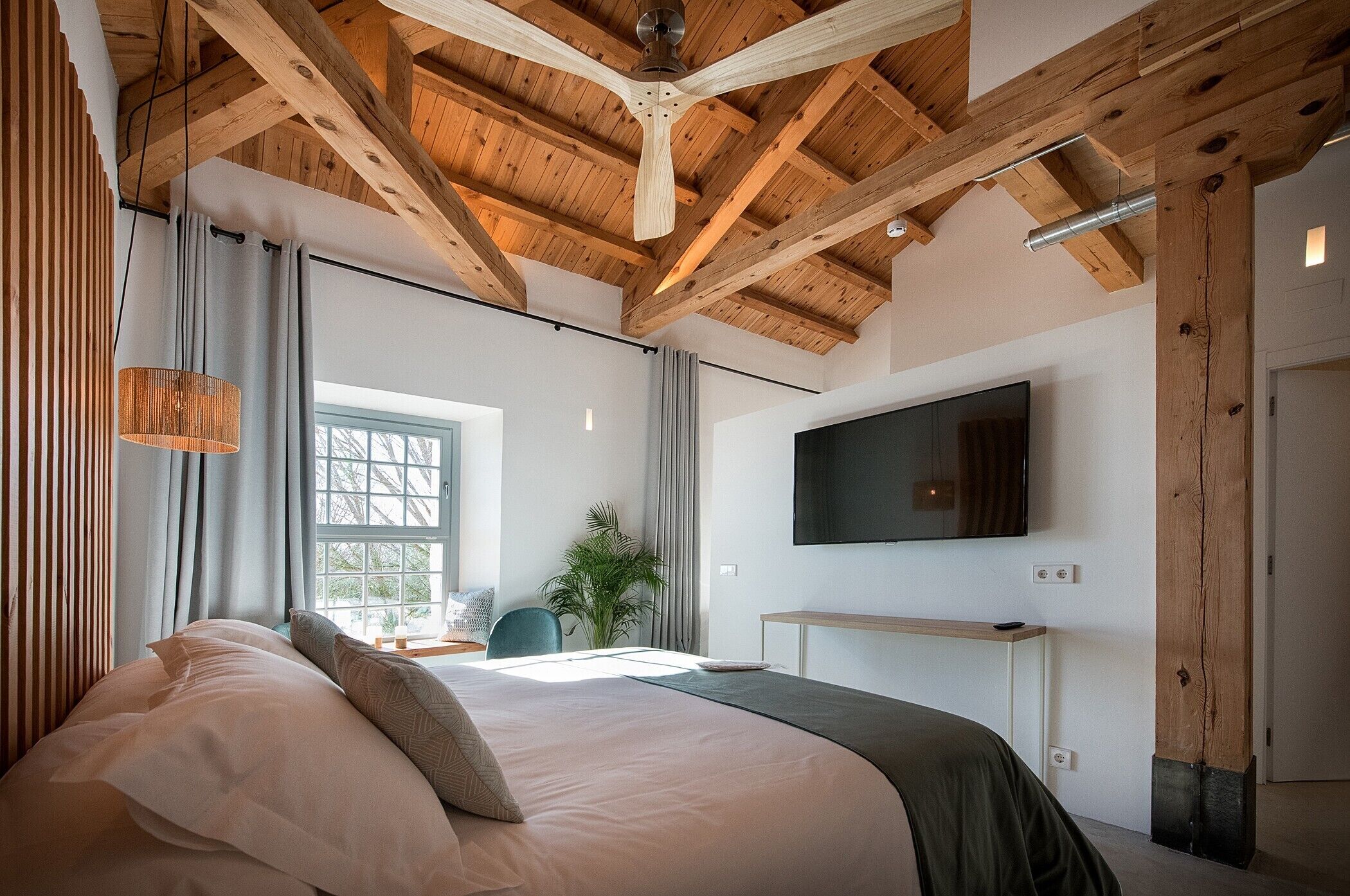 Un dormitorio con una cama grande, vigas de madera en el techo, un televisor de pantalla plana y una ventana con cortinas.