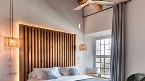 Een moderne slaapkamer met een houten spijbelzijkant, een bed met wit en grijs beddengoed, een bankje, een stoel, en een raam met gordijnen.