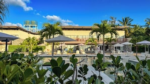 Zona de piscina con sillones de descanso y sombrillas, rodeada de palmeras y un edificio con balcón.