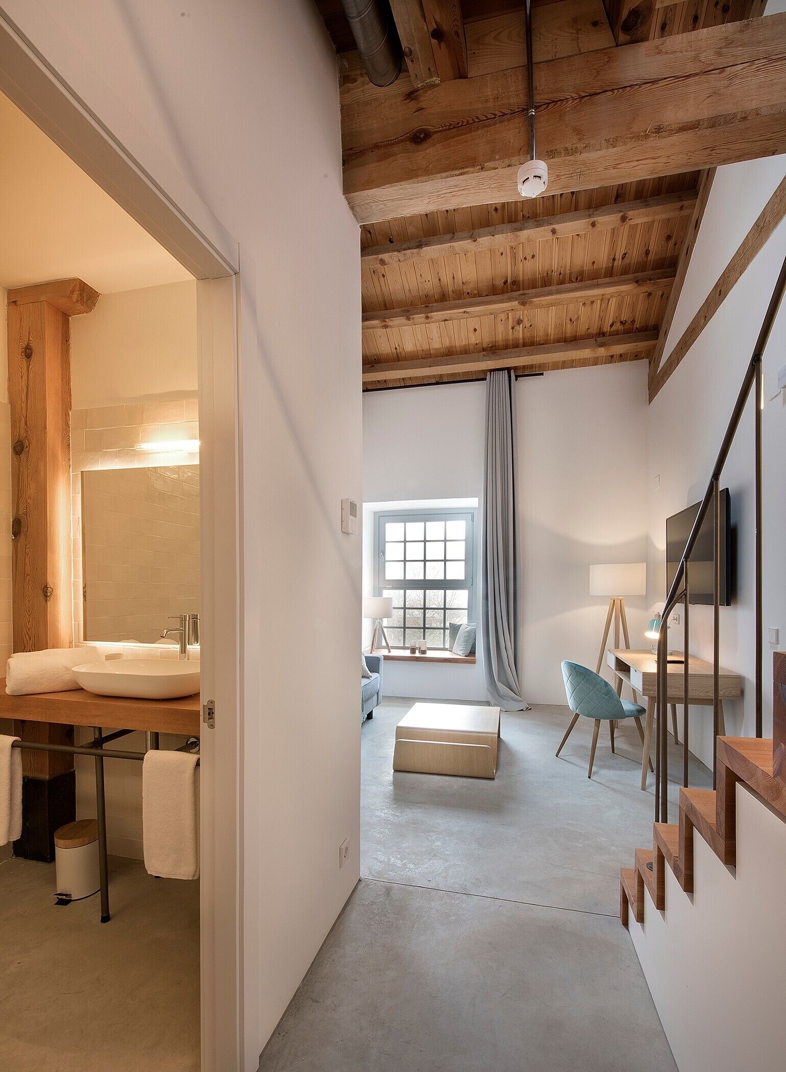 Un intérieur moderne avec un plafond en bois, une salle de bain dotée d’un lavabo et d’un miroir, un coin salon avec une chaise et une table, et un escalier.