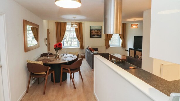 A modern dining area with a round table, chairs, and a pendant light. There is a kitchen area with a stove and a large cylindrical vent. - 32A Bank Street (Rossendale)