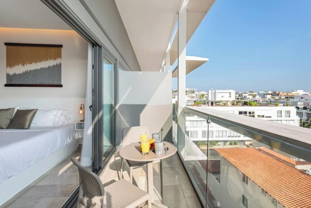 Habitació d'hotel moderna amb un balcó, un llit, una taula petita amb una beguda i vista a la ciutat.