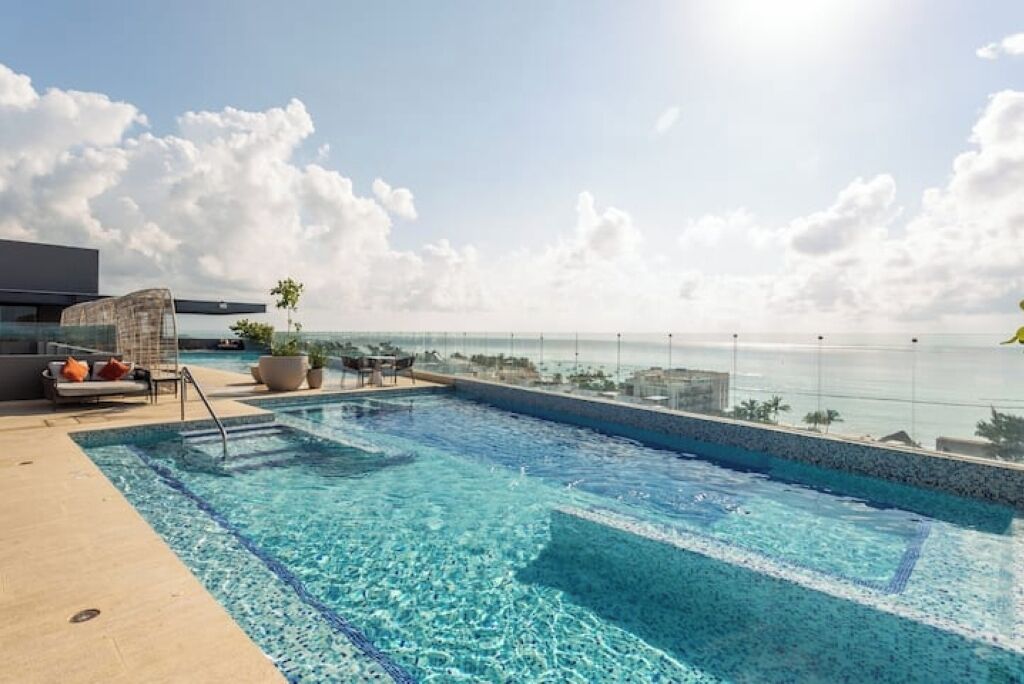 A rooftop infinity pool with clear blue water, a lounge area with a sofa and chairs, and a view of the ocean.