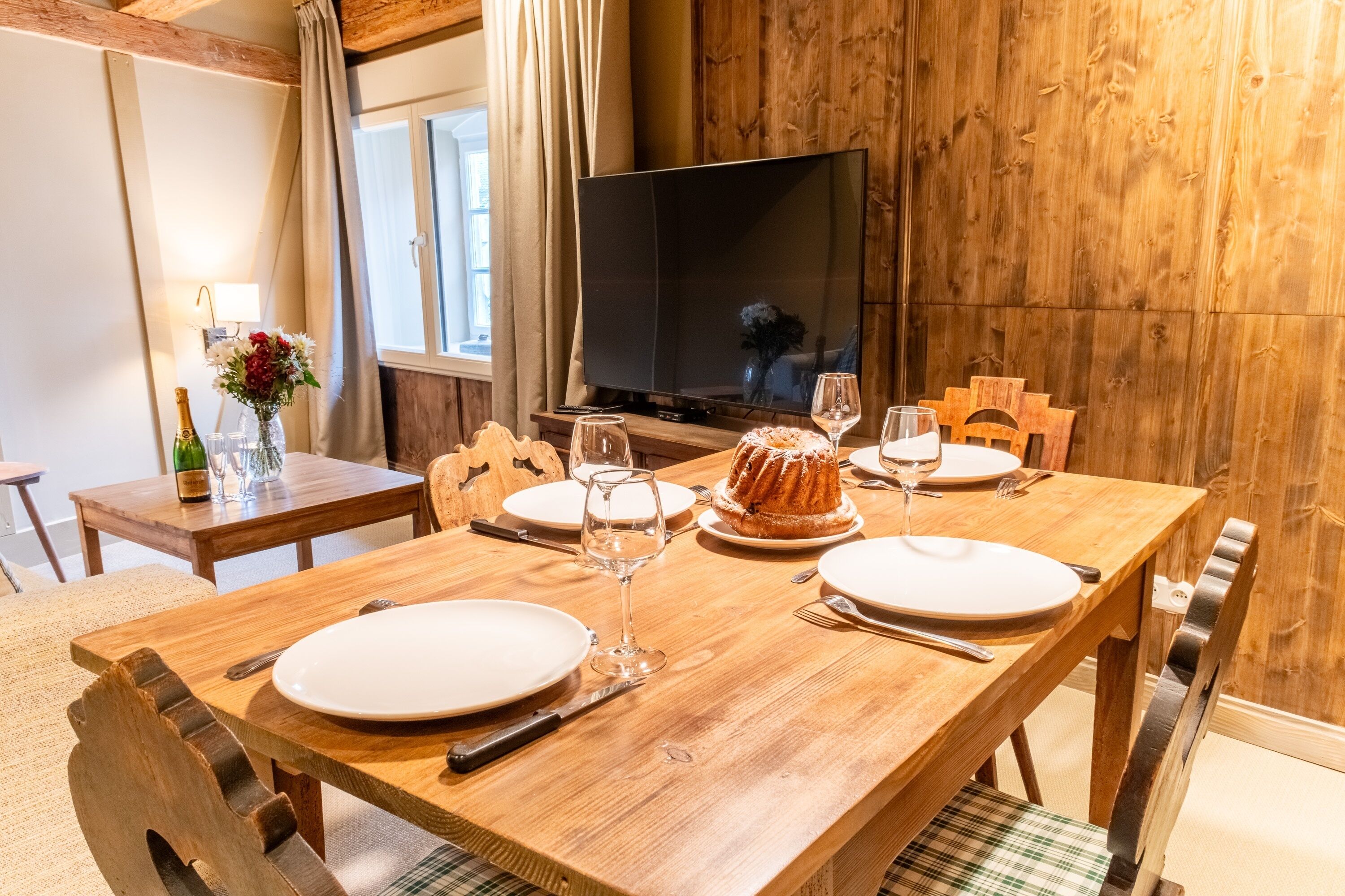 Une table de salle à manger en bois, mise avec des assiettes, des couverts et un gâteau en forme de couronne, avec un téléviseur et un lambris en bois en arrière-plan.