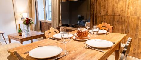 Une table de salle à manger en bois, mise avec des assiettes, des couverts et un gâteau en forme de couronne, avec un téléviseur et un lambris en bois en arrière-plan.