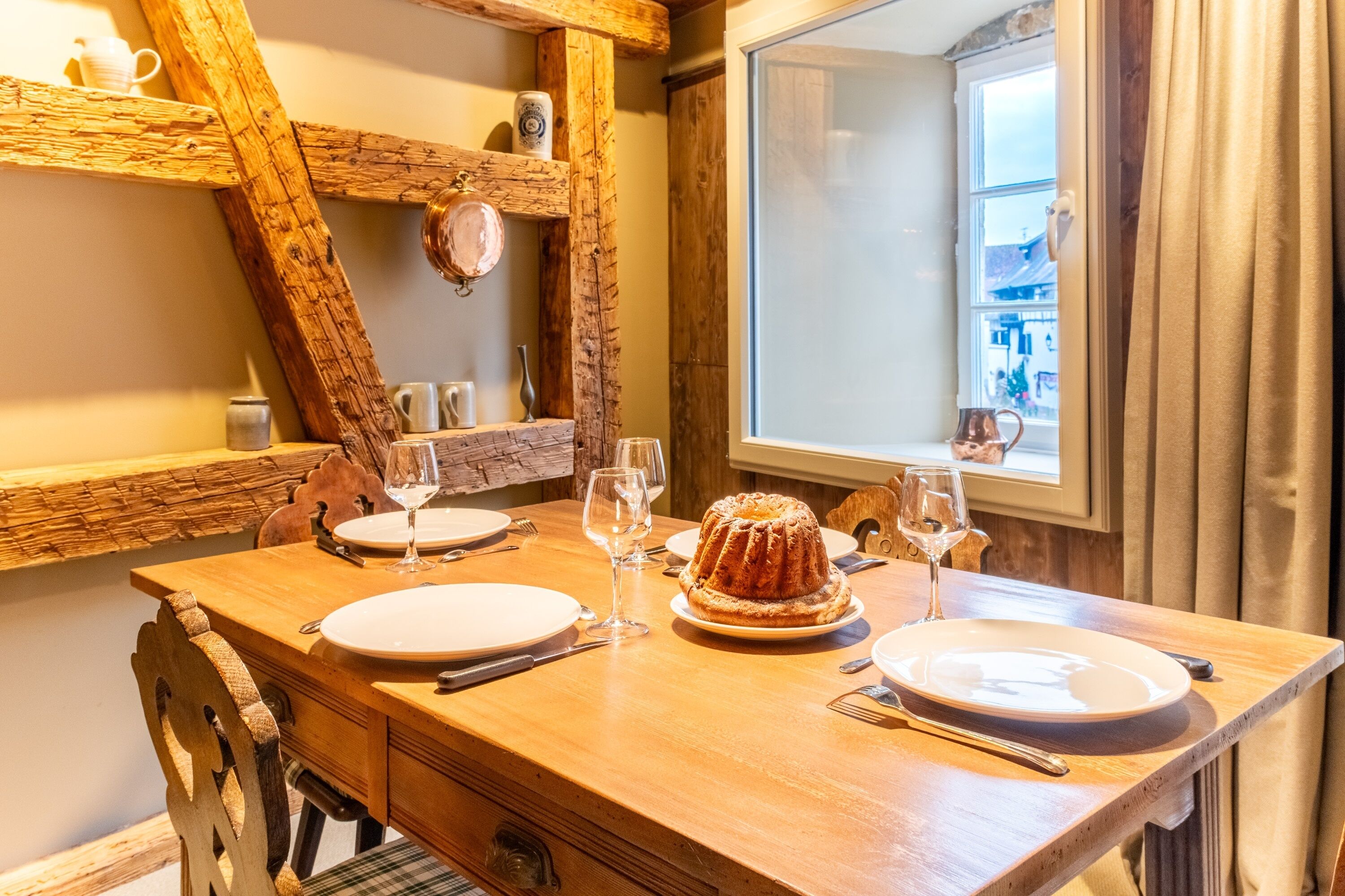 Une table de salle à manger en bois, dressée avec des assiettes, des verres et un gâteau en forme de couronne, avec une fenêtre et des rideaux en arrière-plan.