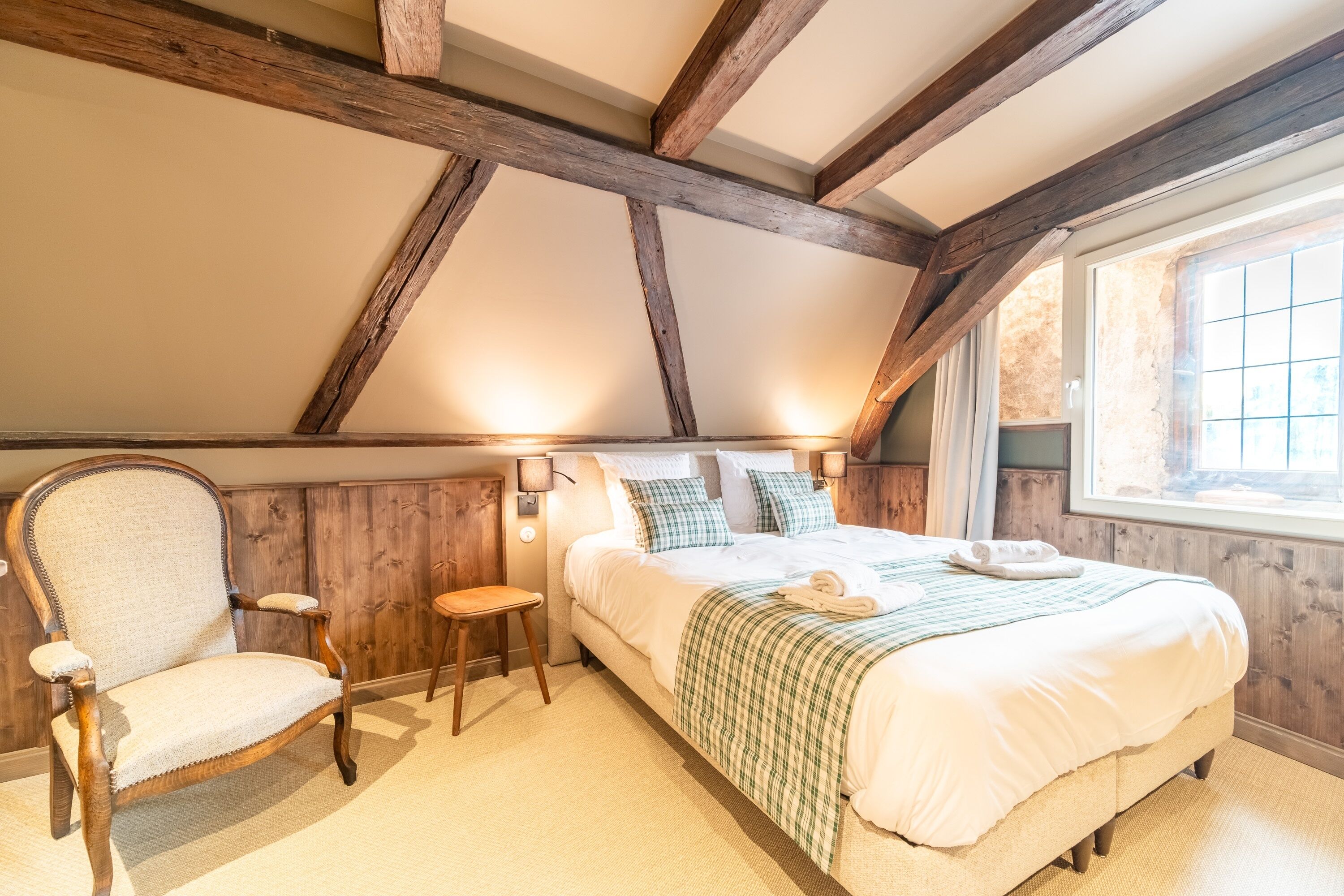 A bedroom with a large bed, wooden beams, a chair, and a window.