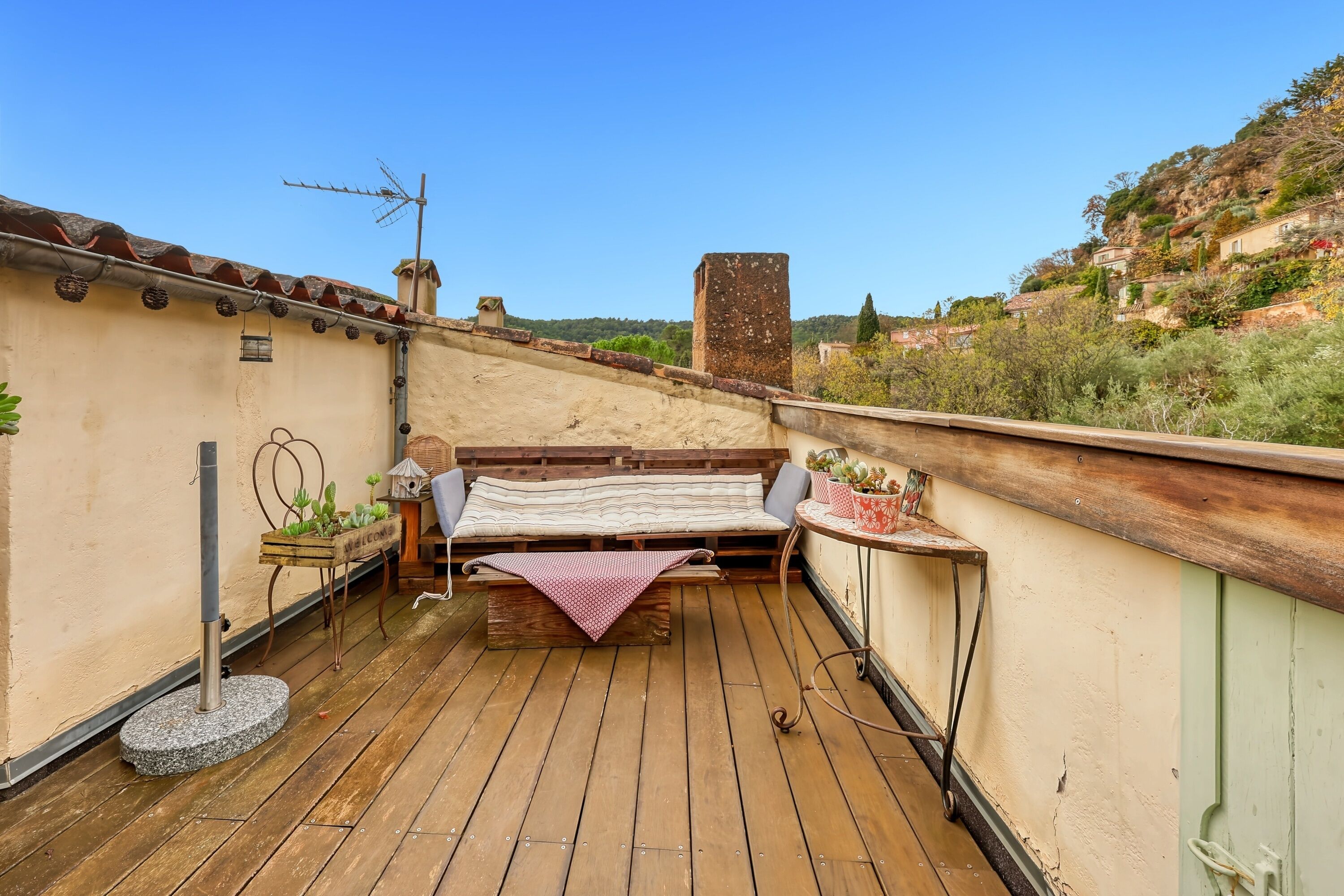 Eine Dachterrasse mit Holzdeck, einer Bank, einem kleinen Tisch mit Topfpflanze und Blick auf eine Hügelkette.