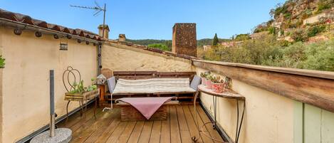 A rooftop terrace with a wooden deck, a bench, a small table with a potted plant, and a view of a hillside.