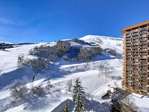 Schneebedeckte Landschaft mit einem modernen Gebäude, Balkonen und einer Skipiste im Hintergrund.