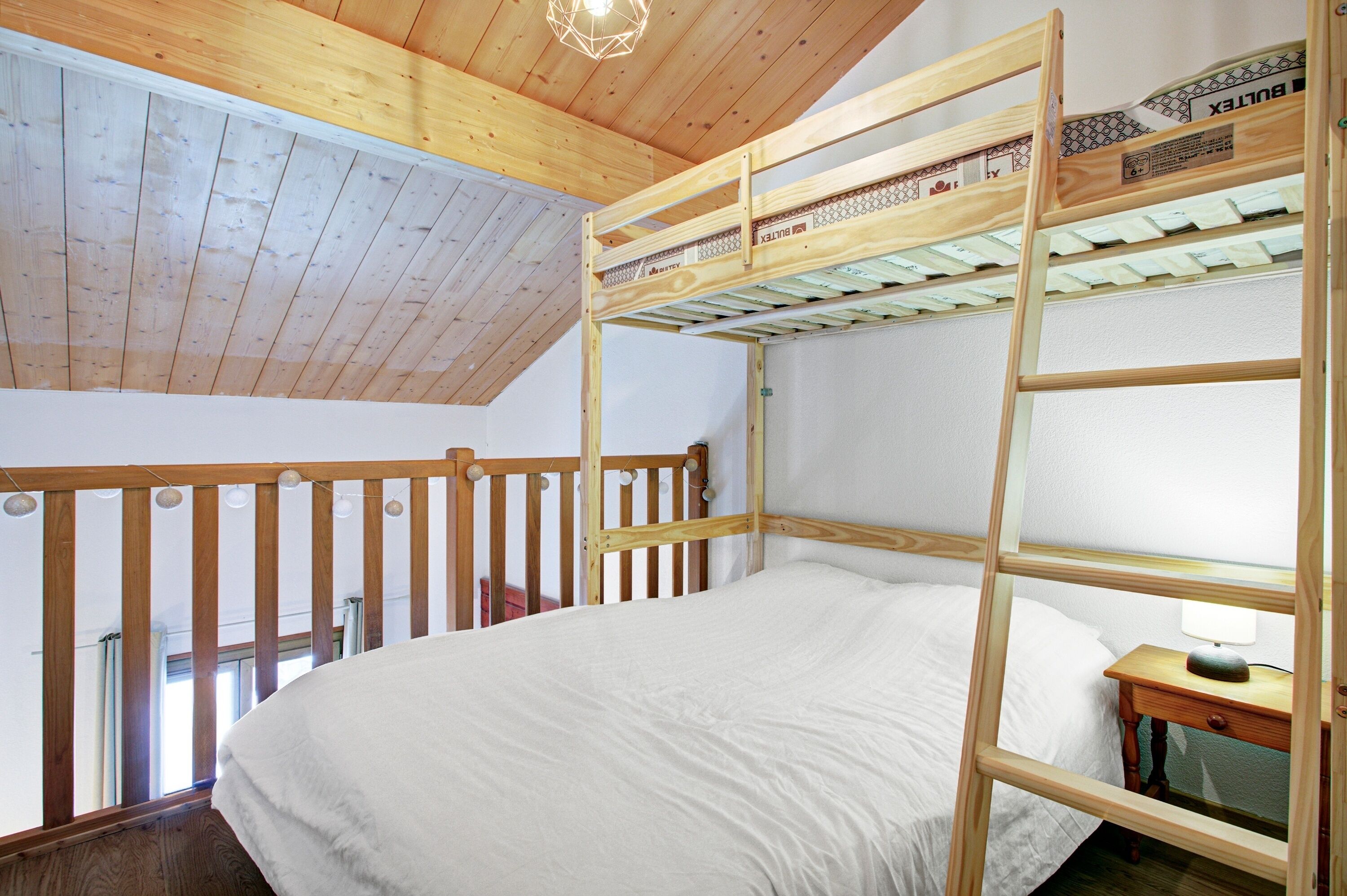 Un lit superposé avec une structure en bois et un matelas blanc, dans une pièce dont le plafond est orné de poutres en bois et qui possède une fenêtre.