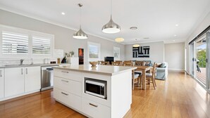 Una cocina moderna con armarios blancos, horno empotrado y una isla central. Hay un comedor con sillas de madera y una mesa, y un ventanal que ofrece vistas al exterior.