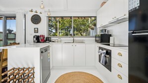 Eine moderne Küche mit weißen Schränken, einem eingebauten Geschirrspüler, einer Mikrowelle und einem Kühlschrank. Ein Fenster bietet Blick auf Bäume, und an der Wand hängt eine Uhr.