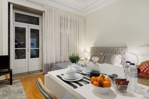 Ein Hotelzimmer mit Essbereich, einem Bett mit einem geblatteten Kopfteil, einem Fernseher und einem Fenster mit Vorhängen.