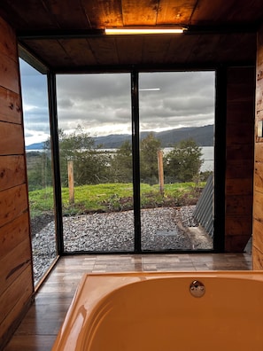 Un interior de madera con un ventanal amplio, una bañera y vistas a un lago y montañas.