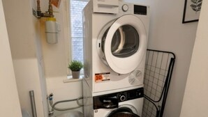 Ein kompakter Waschraum mit einer Waschmaschine und einem Trockner mit Frontlader, einem kleinen Fenster mit Pflanze und einem an der Wand montierten Warmwasserspeicher.