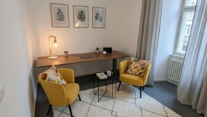 Ein kleiner, moderner Raum mit Schreibtisch, zwei gelben Stühlen, einer Lampe und gerahmten Blätter-Kunstwerken an der Wand.