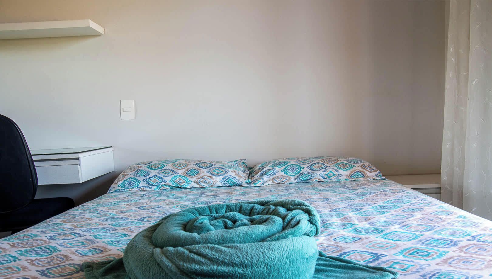 A single bed with patterned bedding, a desk with a chair, and a power outlet on the wall.