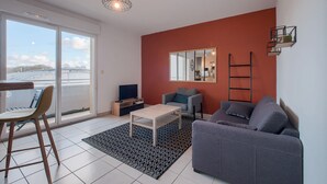 A modern living room with a sofa, a coffee table, a TV, and a large window offering a view of the city. - La Grange - Proche Centre & Parking (Laval)