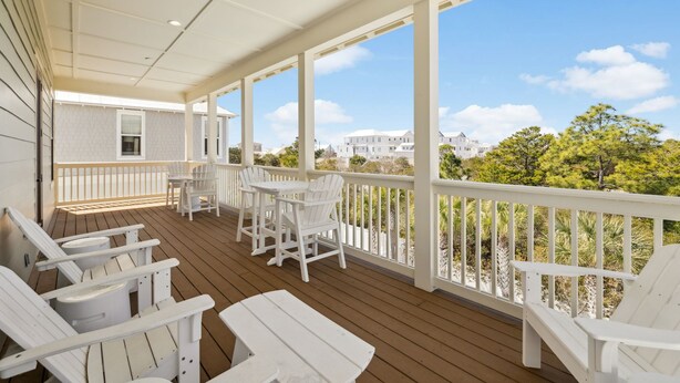 A covered porch with white Adirondack chairs and a table, overlooking a landscaped area with trees and a building in the background. - Pelican Perch (Seagrove Beach)