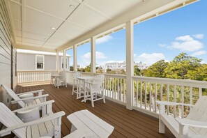 Un porche cubierto con sillas Adirondack blancas y una mesa, con vista a un área ajardinada con árboles y un edificio al fondo.