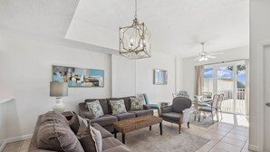 A living room with a sofa, coffee table, and dining area with chairs and a table. There is a ceiling fan and a window with a view of the outdoors. - Seabreeze (101B) (Destin)