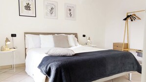 Een net opgemaakt bed met wit beddengoed en een donkerblauwe plaid, een nachtkastje met een lamp, en een wandkapstok.