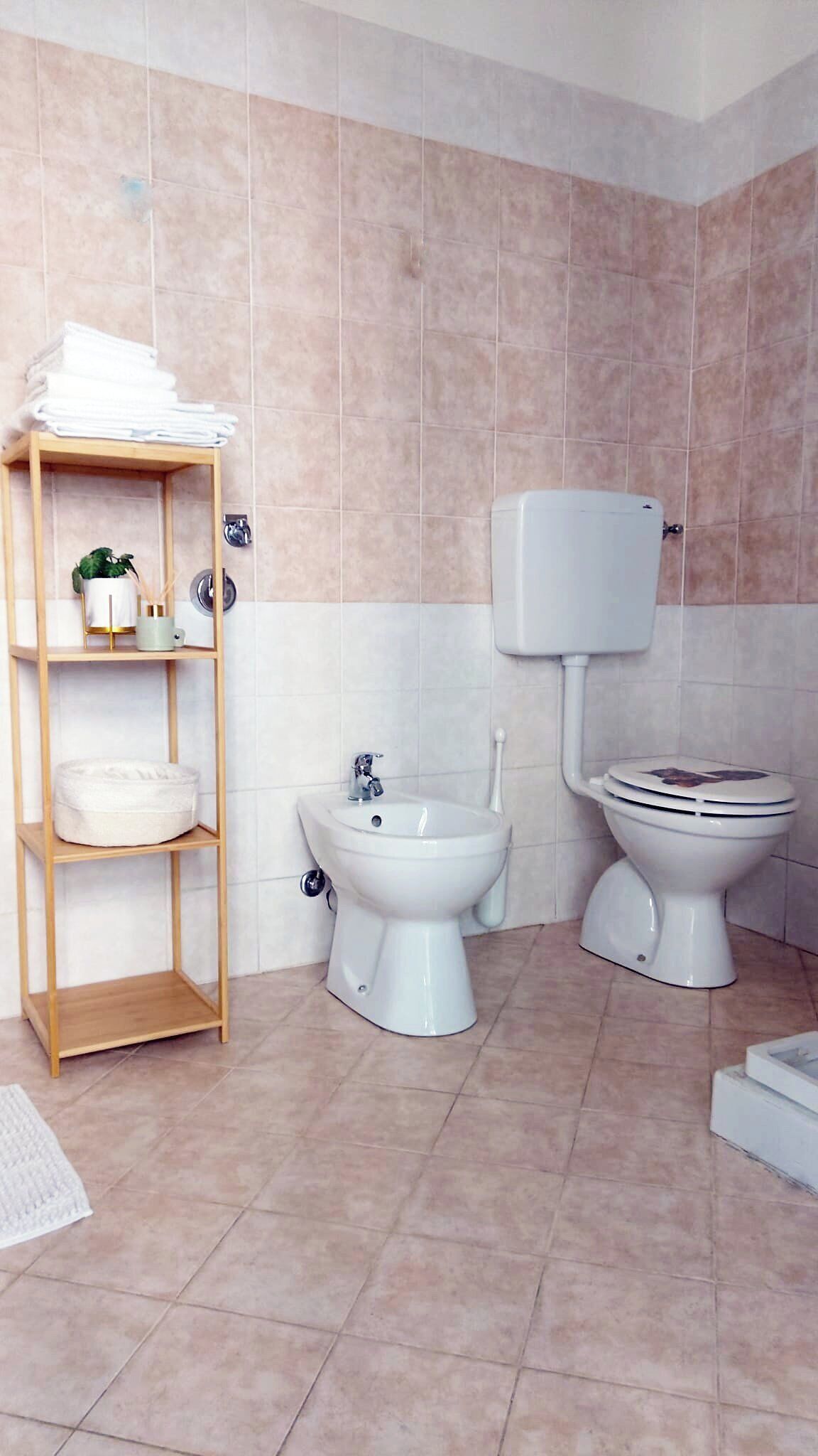 Un baño con inodoro, bidé y lavamanos, un estante de madera con toallas y una planta pequeña.