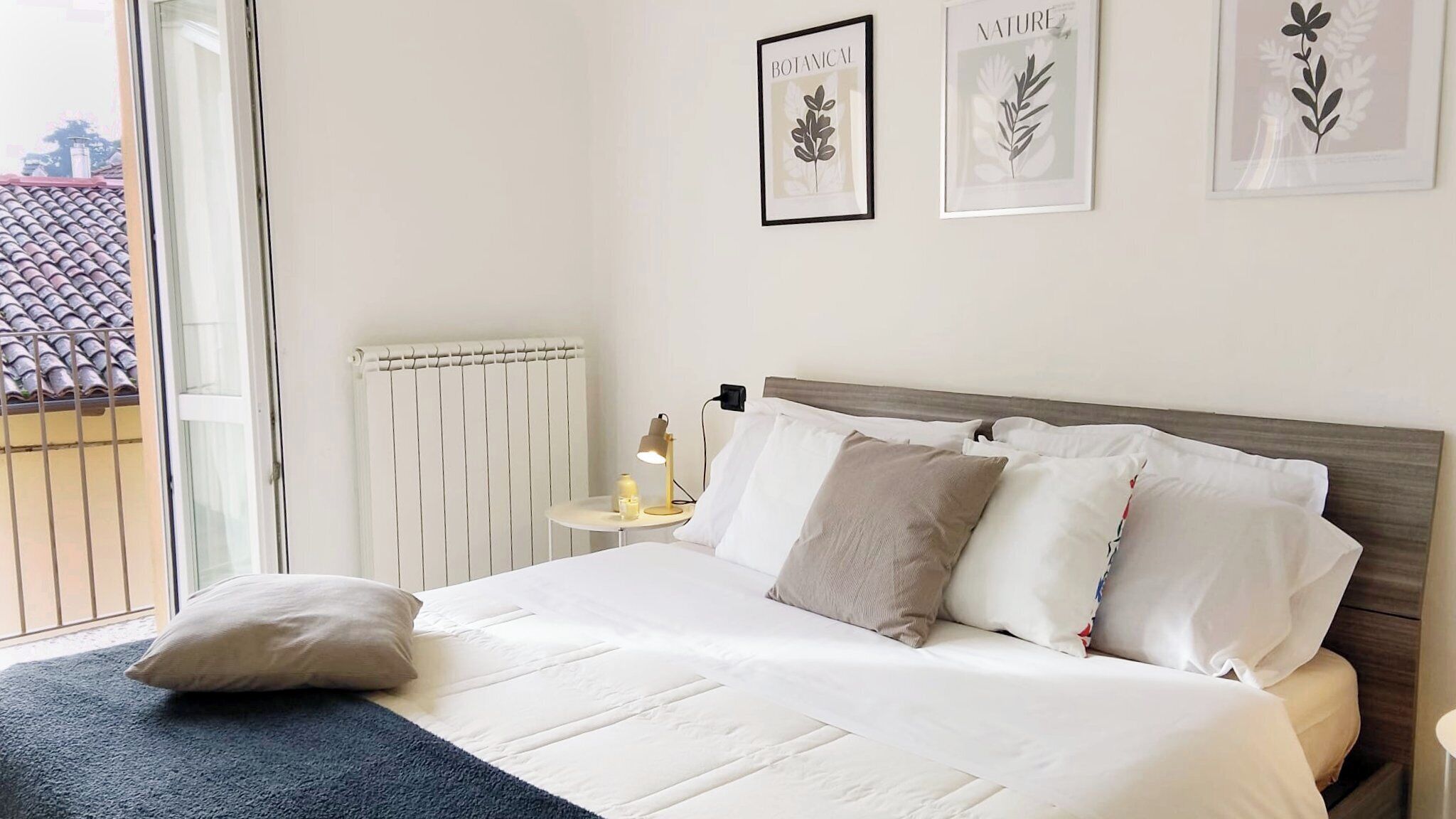 Ein ordentlich bezogenes Bett mit weißen Leinen und mehreren Kissen, ein Nachttisch mit Lampe sowie gerahmte Pflanzenmotive an der Wand.