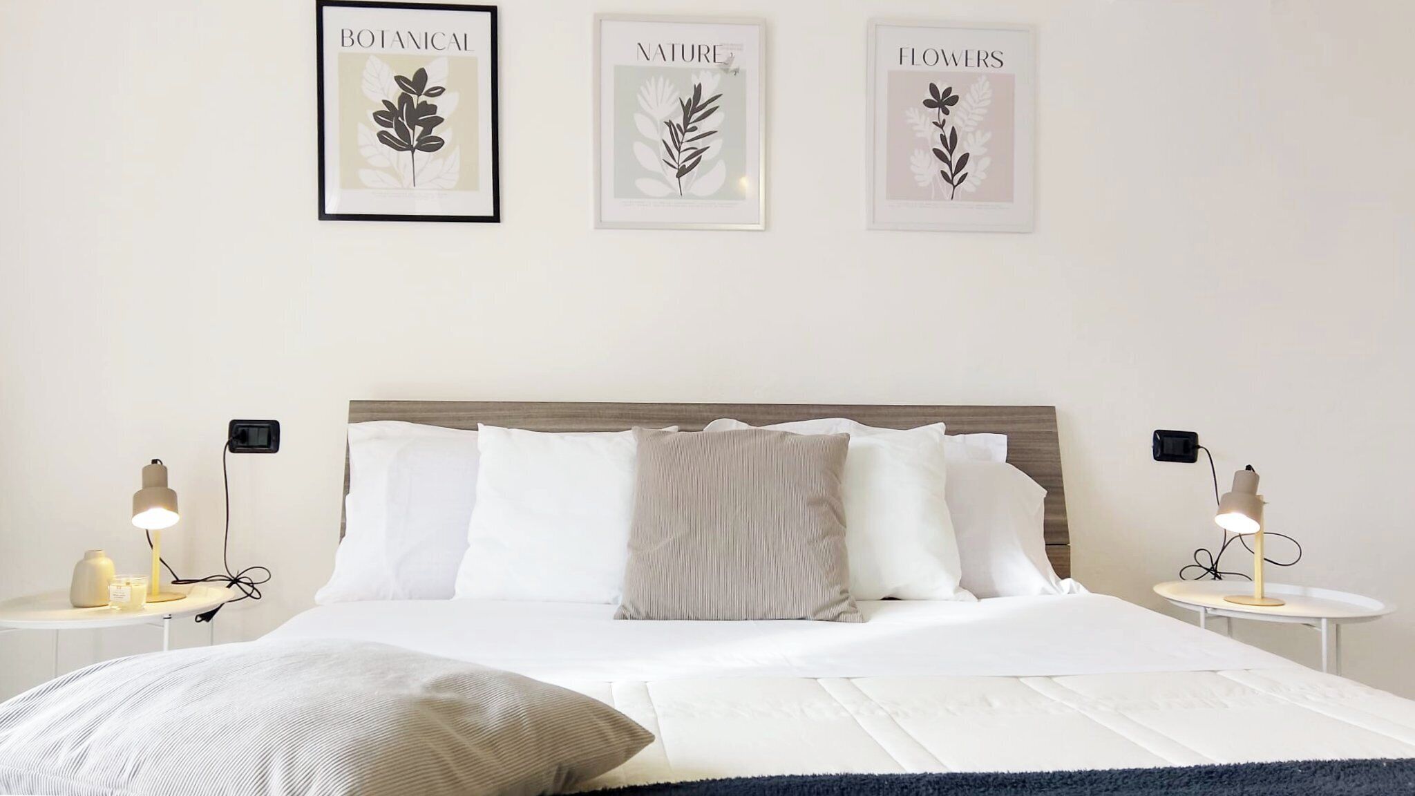 Un lit bien fait, avec du linge de lit blanc, deux tables de chevet avec des lampes et trois reproductions encadrées de motifs botaniques accrochées au mur.