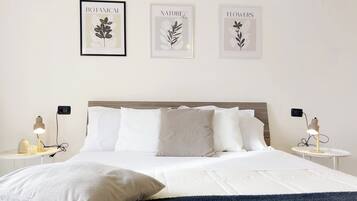 Un lit bien fait, avec du linge de lit blanc, deux tables de chevet avec des lampes et trois reproductions encadrées de motifs botaniques accrochées au mur.