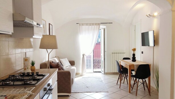Een kleine keuken met een gasfornuis, een eethoek met een tafel en stoelen, een bank en uitzicht op een rode gevel door een raam.