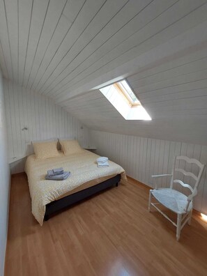 Ein Schlafzimmer mit schrägverdeckter Decke, einem Oberlicht, einem Bett mit gemustertem Überwurf, einem Sessel und Holzböden.