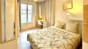 Une chambre à coucher avec un lit, un bureau et une chaise. Il y a une fenêtre avec des rideaux, une porte et un luminaire fixé au mur.