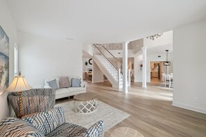 Ein geräumiges Wohnzimmer mit einem weißen Sofa, gemusterten Sesseln und einem runden Couchtisch. Eine Treppe führt auf eine Erschließung.