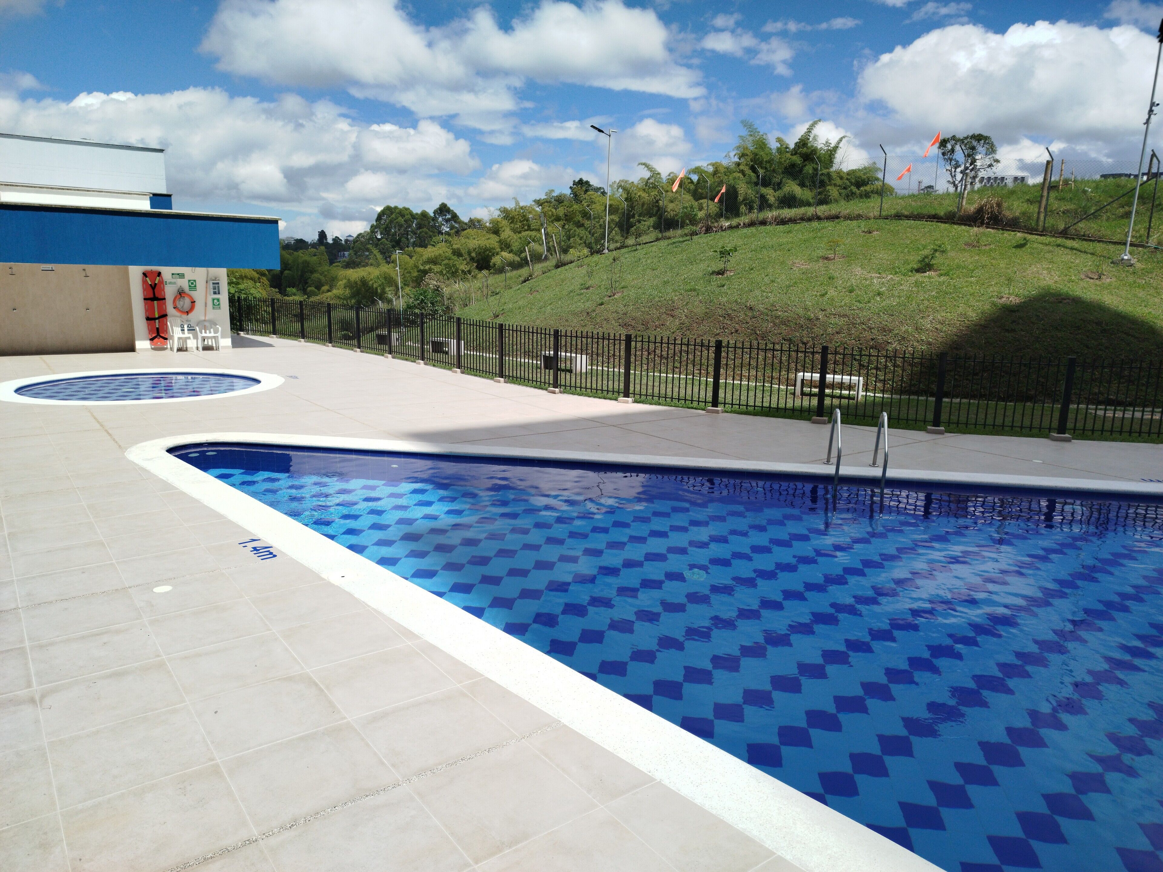 Ein Swimmingpool mit Fliesen umgeben, umzäunt und mit einem grasbewachsen Hügel im Hintergrund.