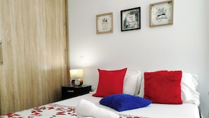 Una cama bien tendida con ropa de cama floral, una toalla blanca y almohadas coloridas. Hay dos cuadros en la pared, uno con un motivo natural y el otro con una cita. Se ve una puerta de madera y una mesita de noche con una lámpara.