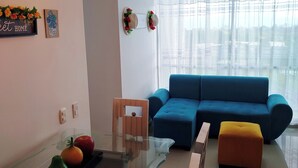 Una sala de estar moderna con un sofá azul, una mesa de centro de vidrio y un reposapiés amarillo. Hay un letrero decorativo en la pared y una ventana con cortinas traslúcidas.