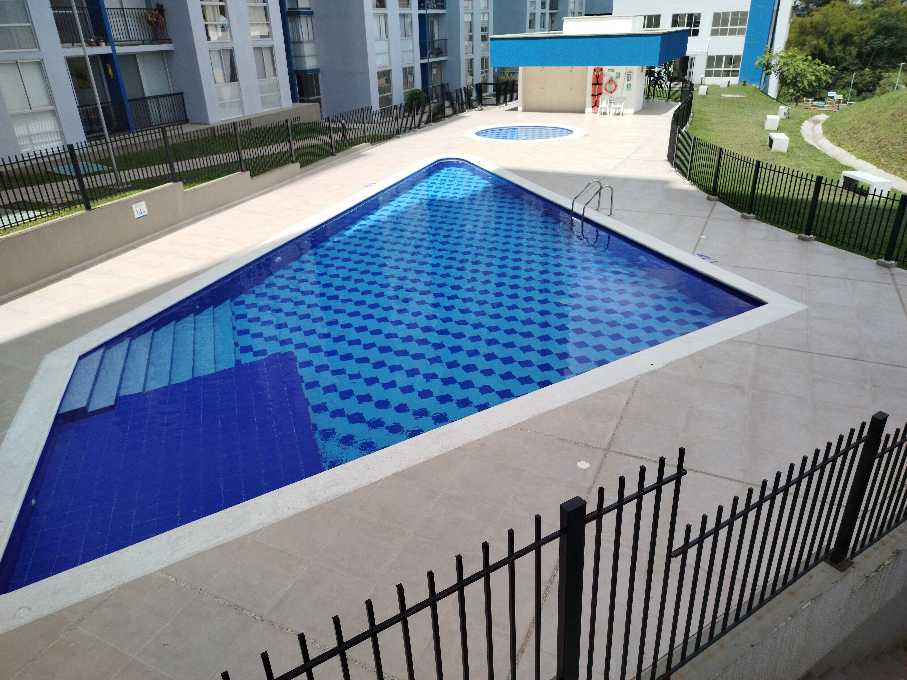 Ein Swimmingpool mit blauem Fliesenmuster, umgeben von einem schwarzen Metallzaun, neben einem Mehrstöckiger Gebäude mit Balkonen.