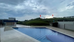 Una piscina con borde de baldosas, rodeada por una valla y una colina cubierta de césped al fondo.