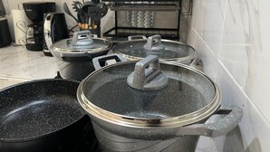 Μια κουζίνα με μαύρες κατσαρόλες στον πάγκο, μια καφετιέρα και μια σχάρα με διάφορα μαγειρικά σκεύη.