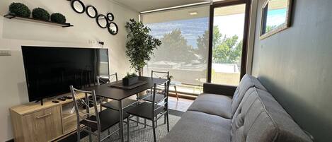 A modern living room with a flat-screen TV, dining table, chairs, sofa, and a large window offering a view of the outdoors.