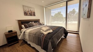 Un dormitorio moderno con una cama grande, una mesita de noche y una ventana que ofrece vistas a árboles y edificios.