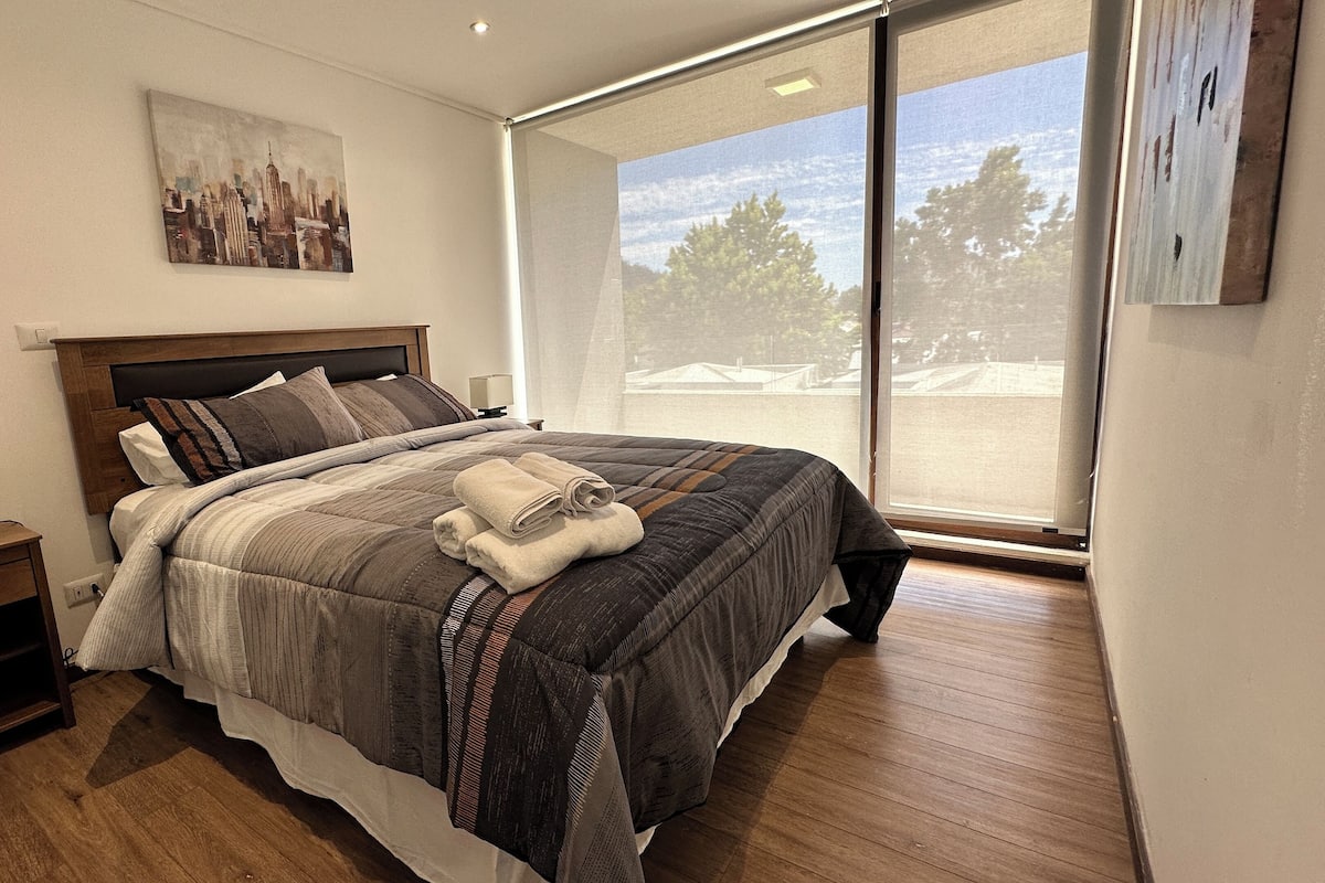 A modern bedroom with a large bed, a nightstand, a bedside table, and a window with a view of trees and buildings.