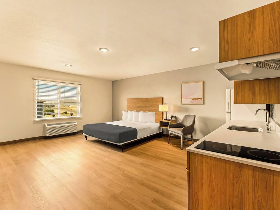 A hotel room with a bed, a desk, a chair, a window with blinds, a radiator, and a kitchenette with a stove.
