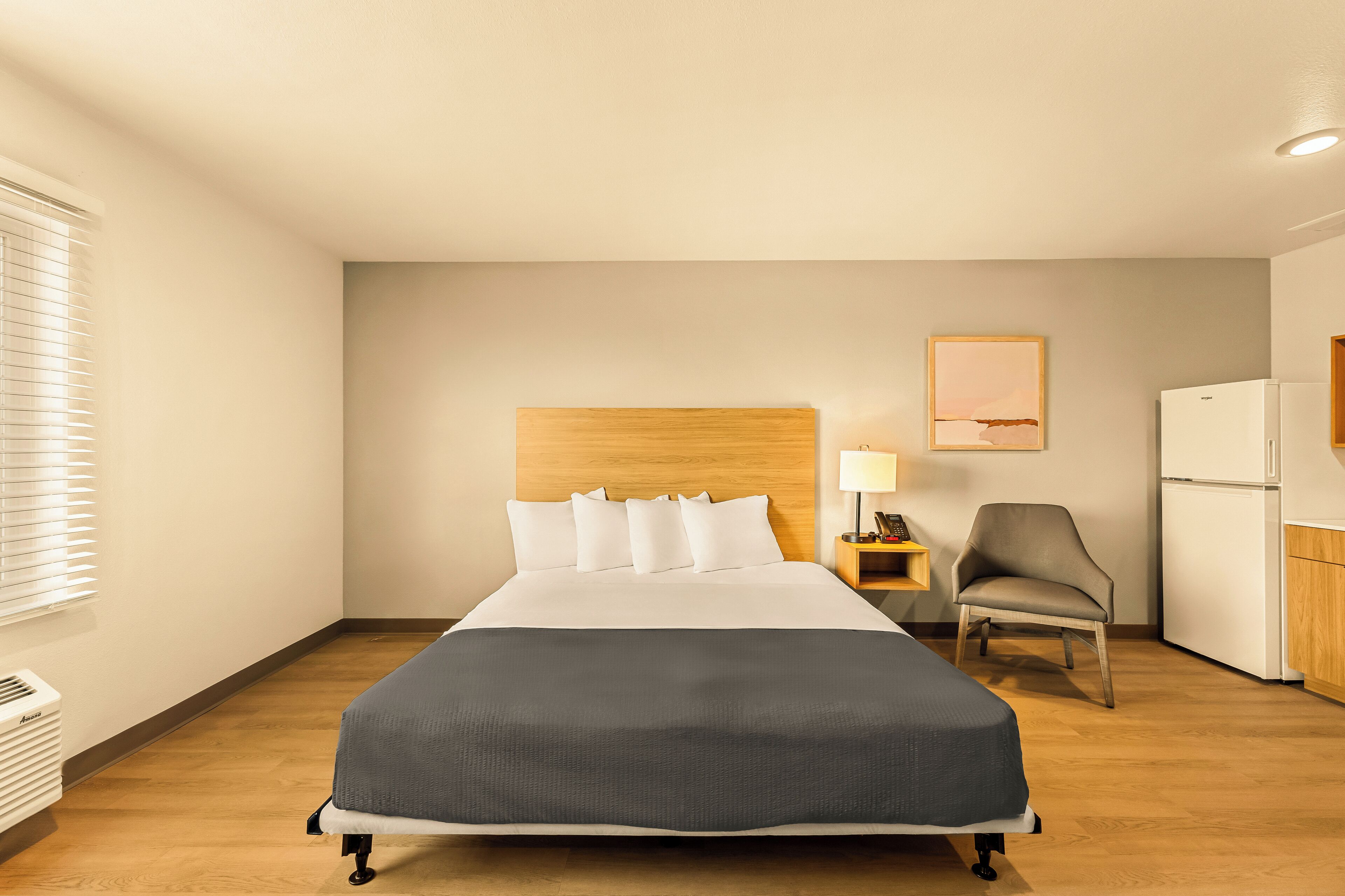 A hotel room with a large bed, a wooden headboard, a desk with a lamp, a chair, and a refrigerator.