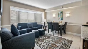 Ein Wohnzimmer mit einer Couch, einem Hocker und einem Teppich mit Muster. Es gibt einen Essbereich mit Stühlen und einem Tisch.