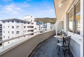 En balkon med et bord og stole, med udsigt over et boligområde med bygninger og en bakke i baggrunden.