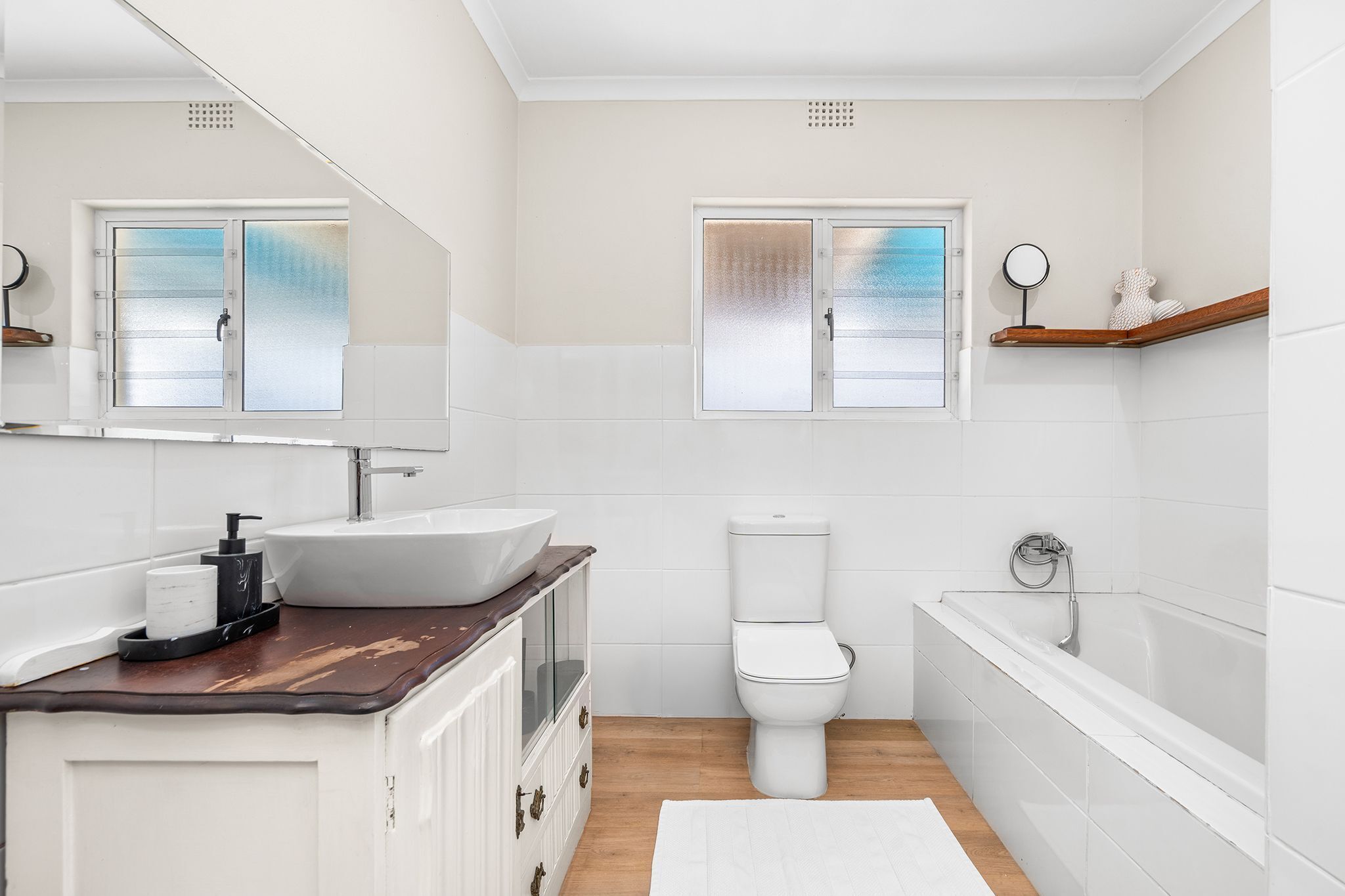 Un baño con lavamanos, inodoro y bañera blancos, tocador de madera y ventanas con vidrio esmerilado.