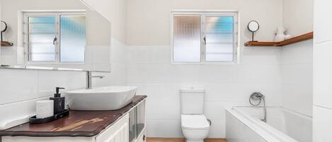 Un baño con lavamanos, inodoro y bañera blancos, tocador de madera y ventanas con vidrio esmerilado.